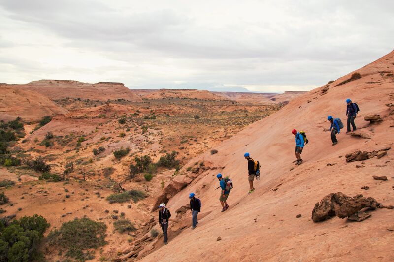 A group of hikers is traversing a vast, arid landscape. They are walking single file up a reddish-orange rock formation, with backpacks indicating they are on a trek. The background reveals a sprawling desert environment under a cloudy sky, suggesting a remote and adventurous journey.